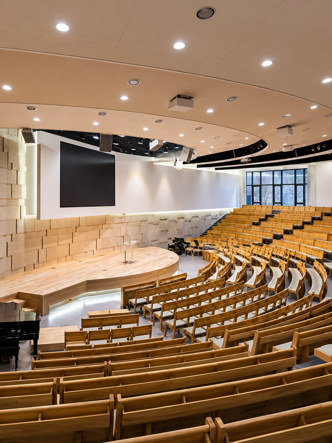 Auditorium with curved rows of tiered wooden seats facing a stage and wall-mounted screen