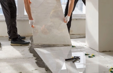 Two workers lowering a large light-colored tile onto floor adhesive