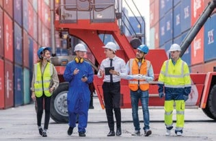 Five workers in hard hats walking beside a red container handler and stacked shipping containers