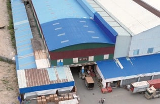 Aerial view of blue-roofed industrial buildings around a loading yard with trucks and crates