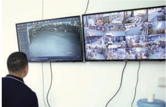 Man viewing two wall-mounted monitors showing a yard camera feed and a grid of warehouse camera views