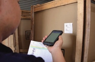 Worker holding a handheld device and paperwork beside a label on a wooden shipping crate