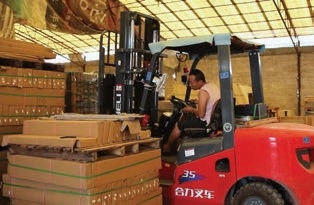 Red forklift operator handling stacked cardboard cartons secured with green straps inside a warehouse